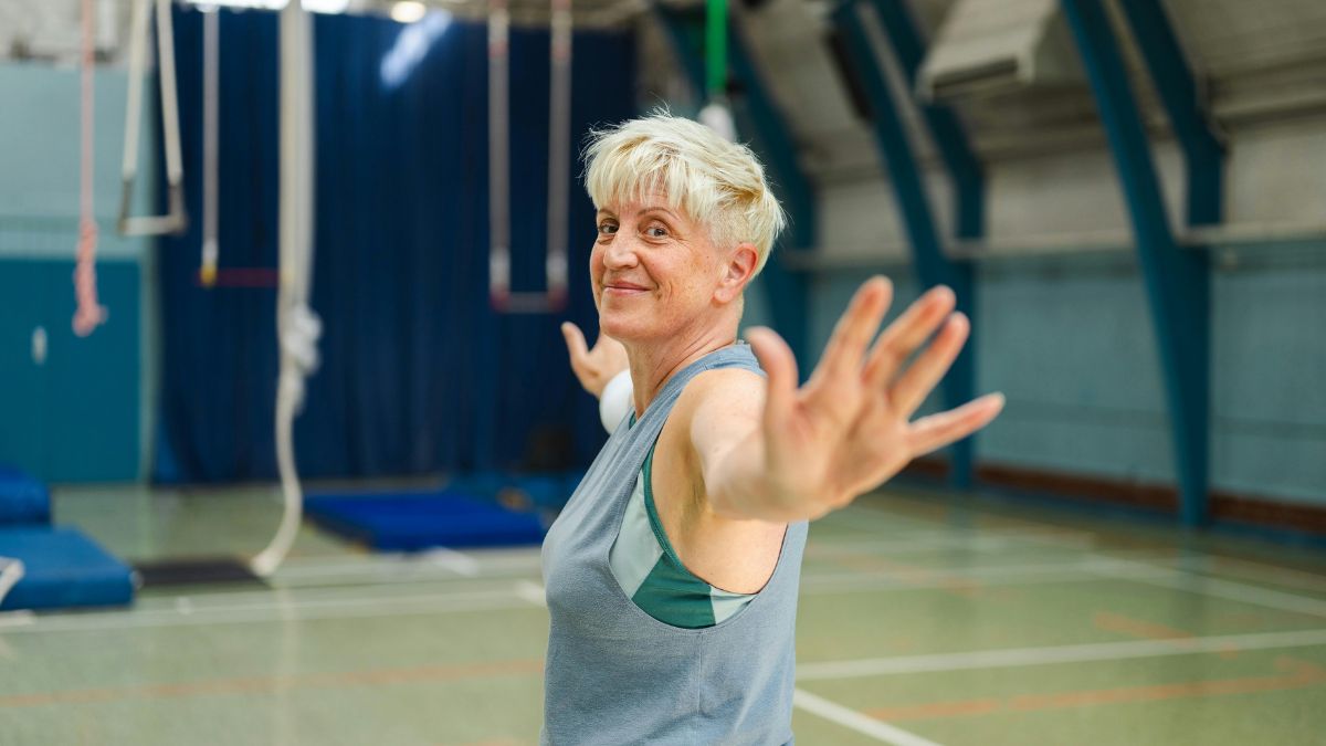 Žena s krátkymi blond vlasmi cvičí v telocvični, usmieva sa a má otvorené ruky.