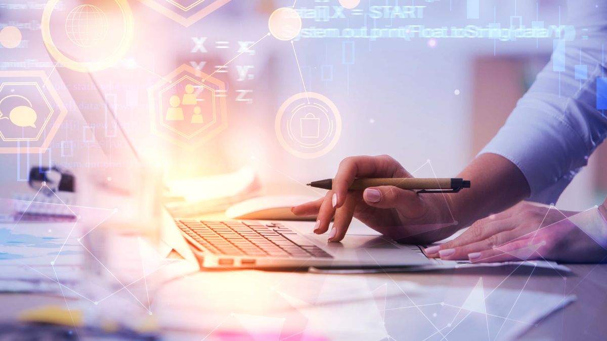 Ruka s perom píše na papier pri počítači s digitálnymi prvkami v pozadí.