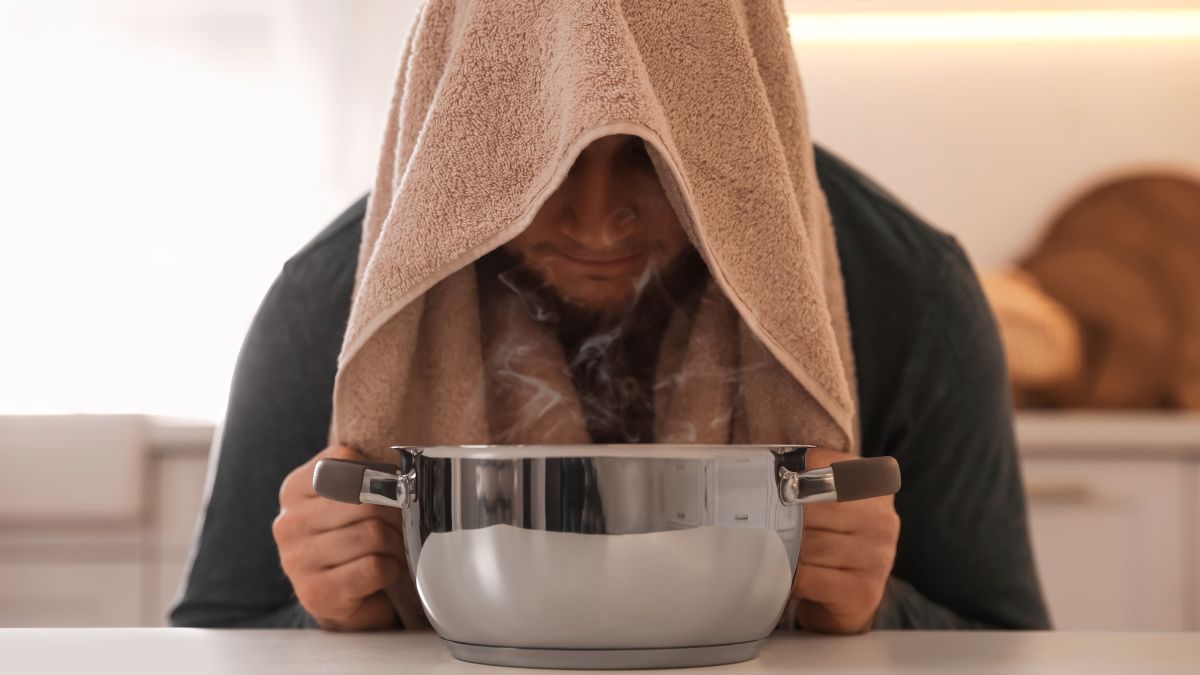 Muž inhaluje paru zo hrnca, zakrytý uterákom, v domácom prostredí.