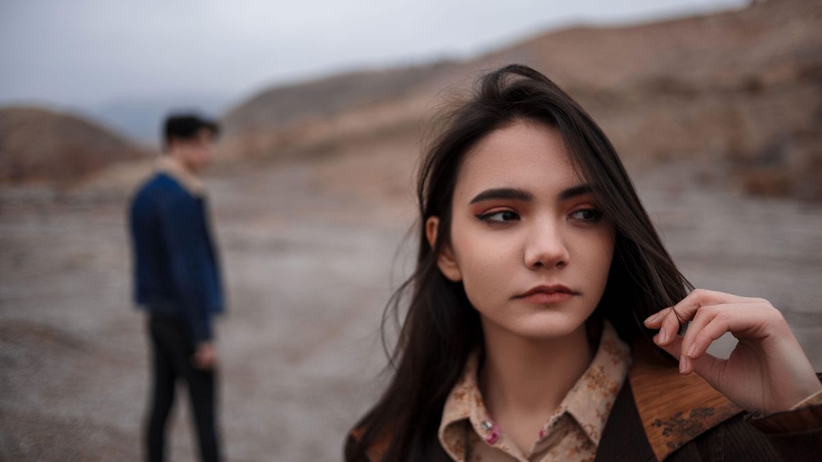 Žena s vážnym výrazom na tvári, pozorujúca okolie, v pozadí muž.