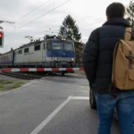 Osoba čaká pred železničnými závorami a prichádza vlak.